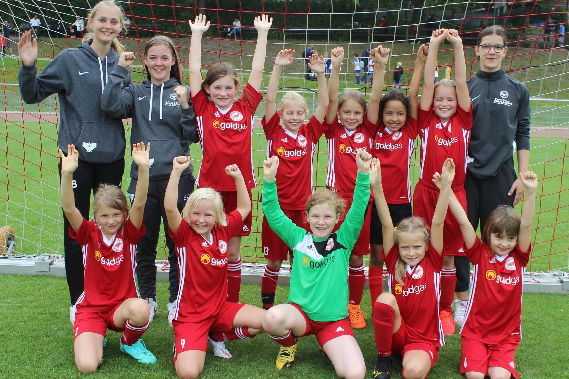 Mädchenfußballteam in roten Trikots posiert fröhlich vor einem Fußballtor, einige Spielerinnen heben die Arme.