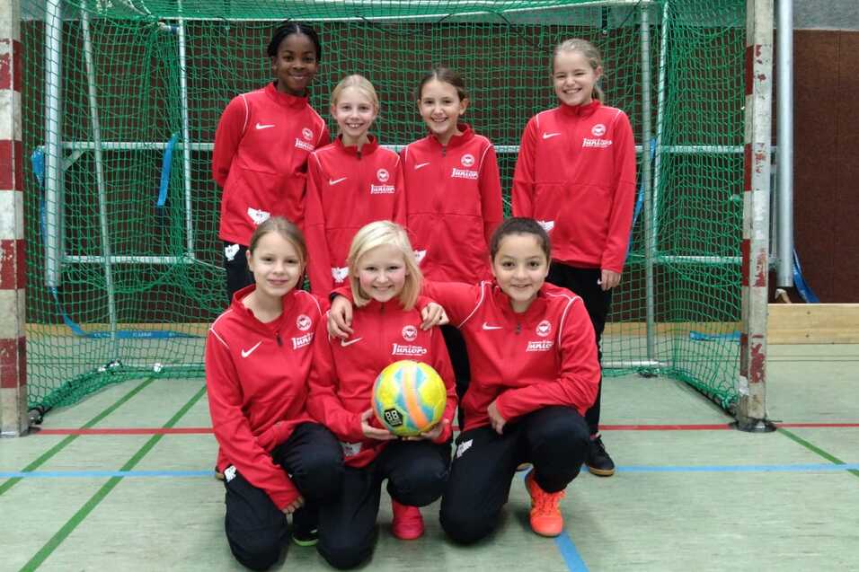 Gruppe von sieben Mädchen in roten Trainingsanzügen, posierend vor einem Handballtor mit einem Ball in der Mitte.