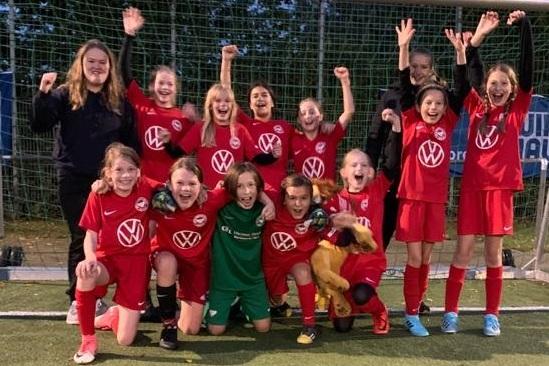 Gruppenselfie von Mädchenfußballspielerinnen in roten Trikots, zufriedene Gesichter, Jubel vor dem Tor.