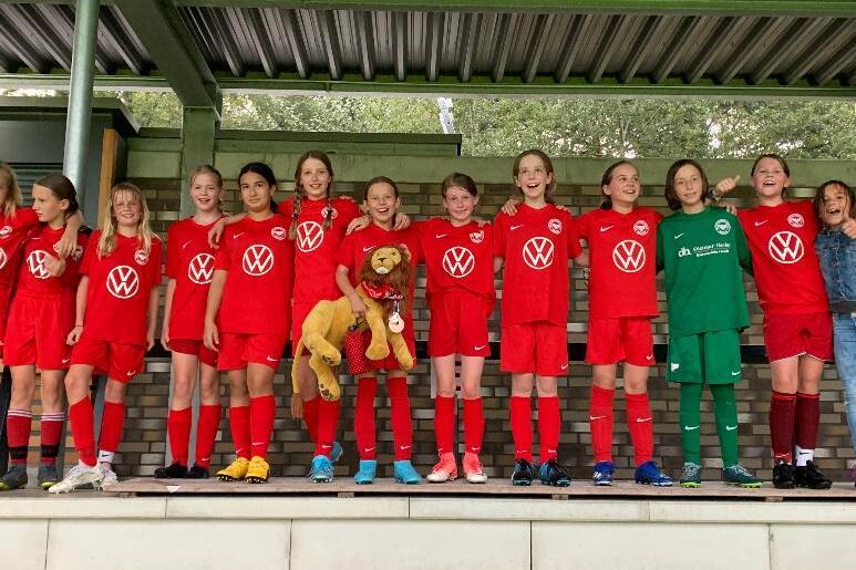 Mädchenfußballmannschaft in roten Trikots mit einem Maskottchen auf einer Tribüne, lächelnd für das Foto aufgestellt.