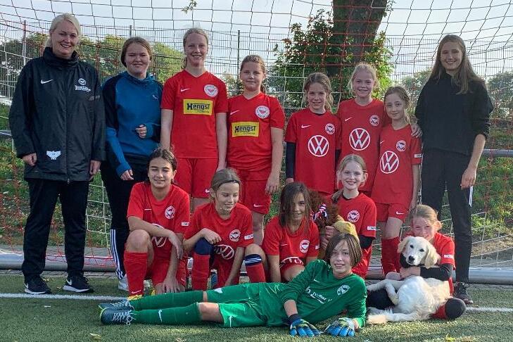 Mädchenfußballmannschaft in roten Trikots posiert mit Trainerin vor einem Fußballtor auf einem Kunstrasenplatz.