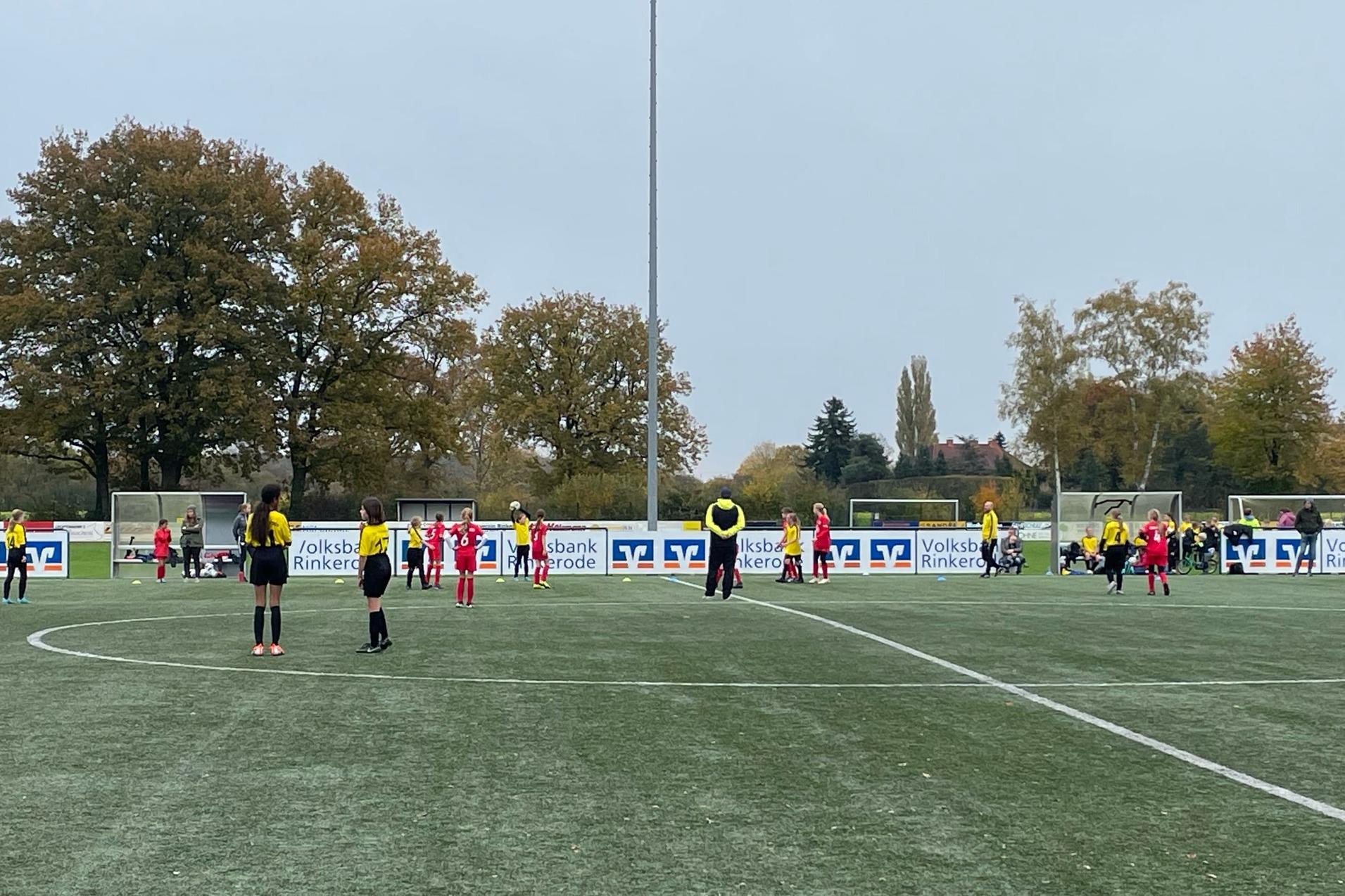Kindermannschaft auf einem künstlichen Fußballplatz, umgeben von Bäumen, spielt unter grauem Himmel.