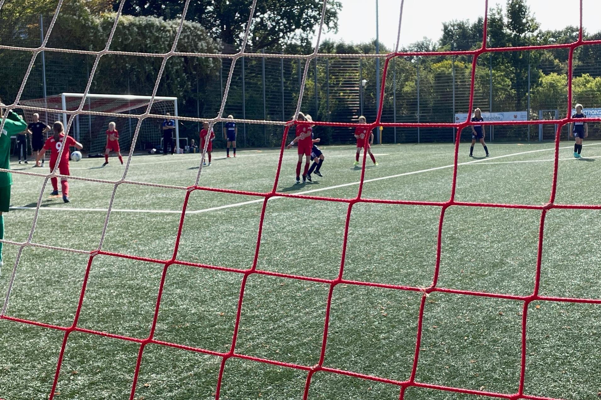 Sicht durch ein Fußballnetz auf ein Spiel, Spieler in roten und blauen Trikots laufen über das grüne Kunstrasenfeld.