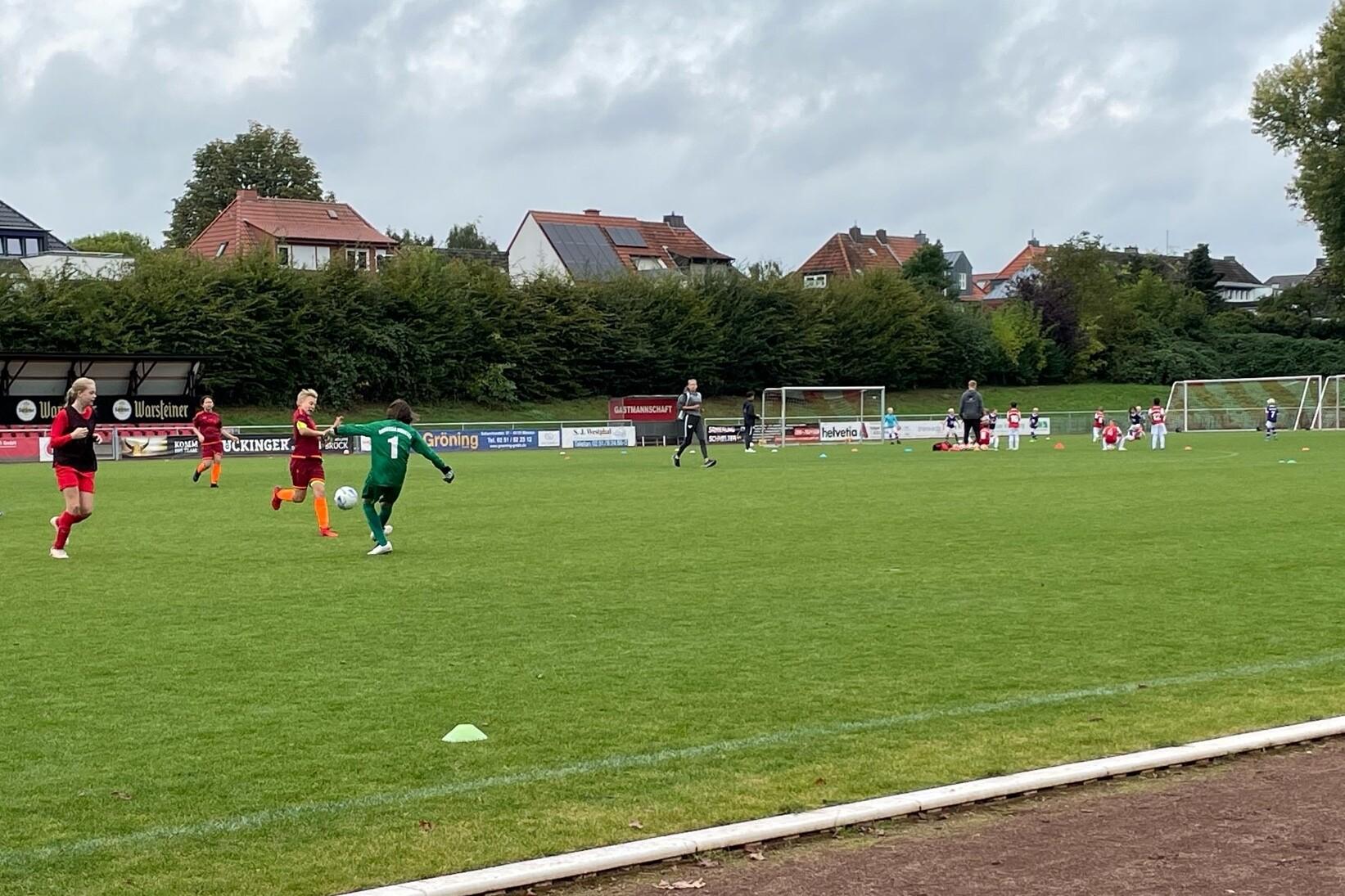 Ein Fußballspiel mit Spielern in roten und grünen Trikots auf einem Rasenplatz, im Hintergrund Tore und Zuschauer.