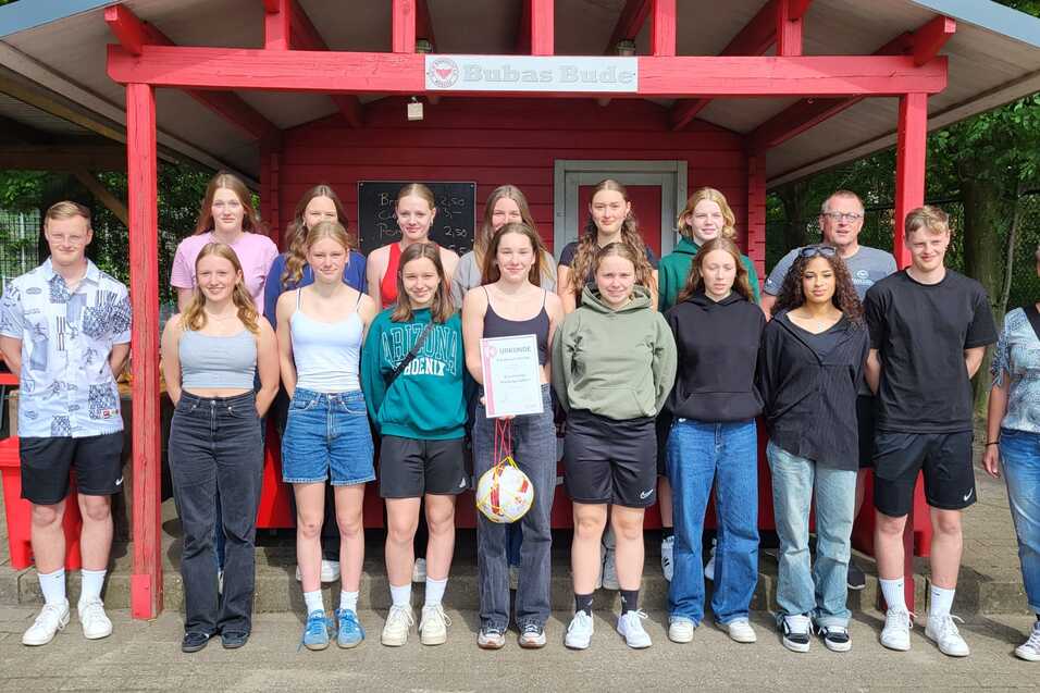 Gruppenfoto von 16 Personen vor einer roten Holzhütte, einige halten einen Volleyball, alle lächeln in die Kamera.