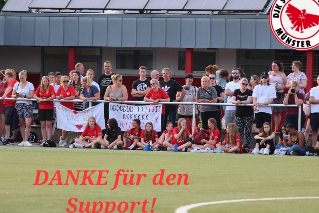 Eine Gruppe von Zuschauern mit roten Trikots steht an einem Fußballfeld, einige halten Banner mit Dankesschriften.