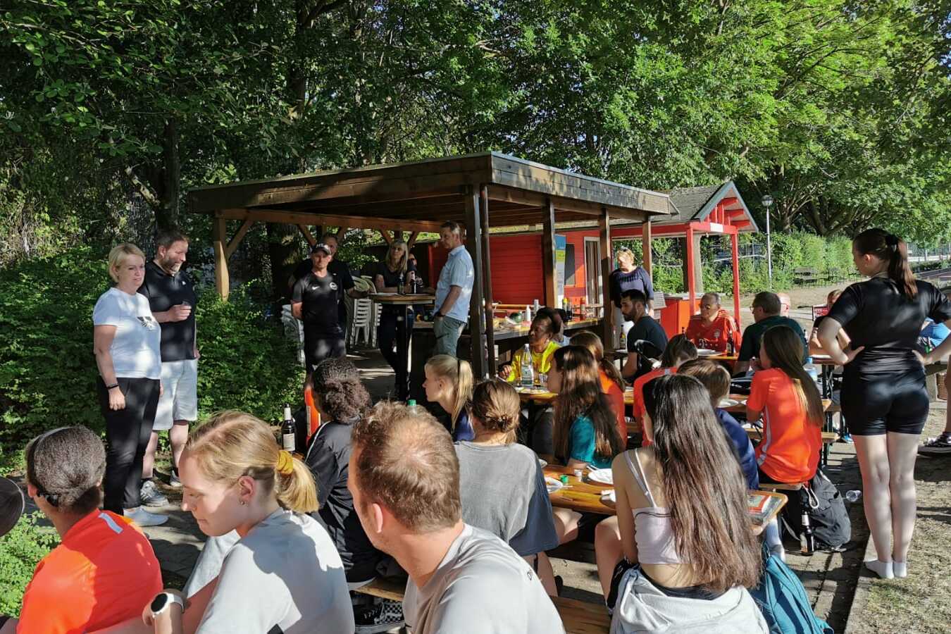 Gruppenszene mit Sportlern, die an Tischen sitzen und essen, umgeben von Grün und einem gemütlichen Kiosk.