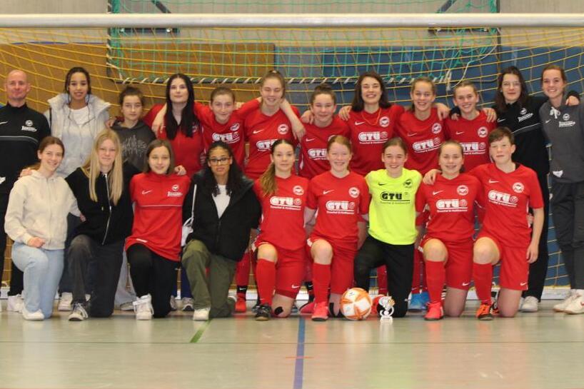 Gruppenfoto einer Fußballmannschaft in roten Trikots, zusammen mit Trainerinnen, in einer Sporthalle vor dem Tor.