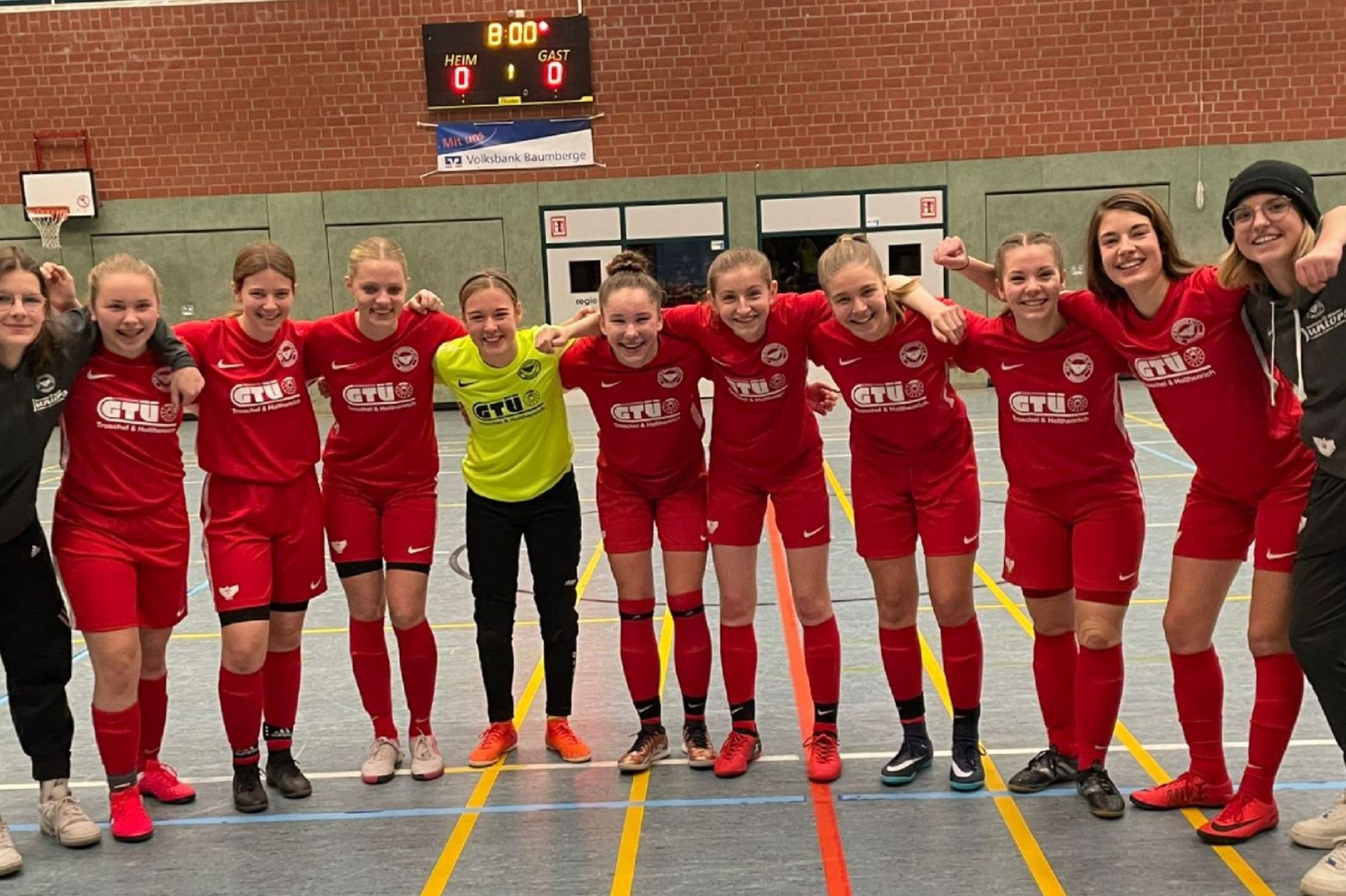 Eine Gruppe von elf Fußballspielerinnen in roten Trikots posiert lächelnd auf einem Hallenfußballfeld.