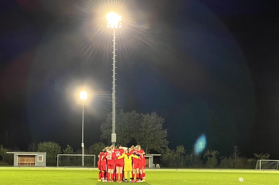 Eine Fußballmannschaft in roten Trikots versammelt sich im nächtlichen Stadion unter Flutlicht auf einem Rasenfeld.