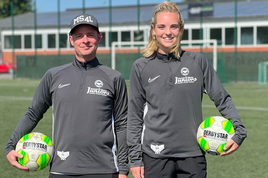 Zwei Personen in Sportkleidung halten jeweils einen Fußball auf einem grünen Fußballfeld.