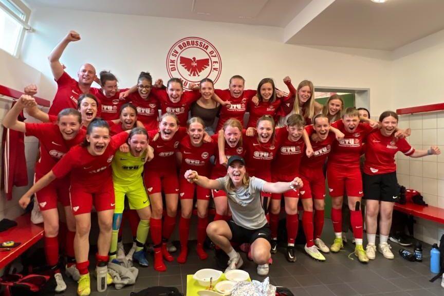 Gruppenfoto von Fußballspielerinnen in roten Trikots, die jubelnd in einem Umkleideraum stehen.