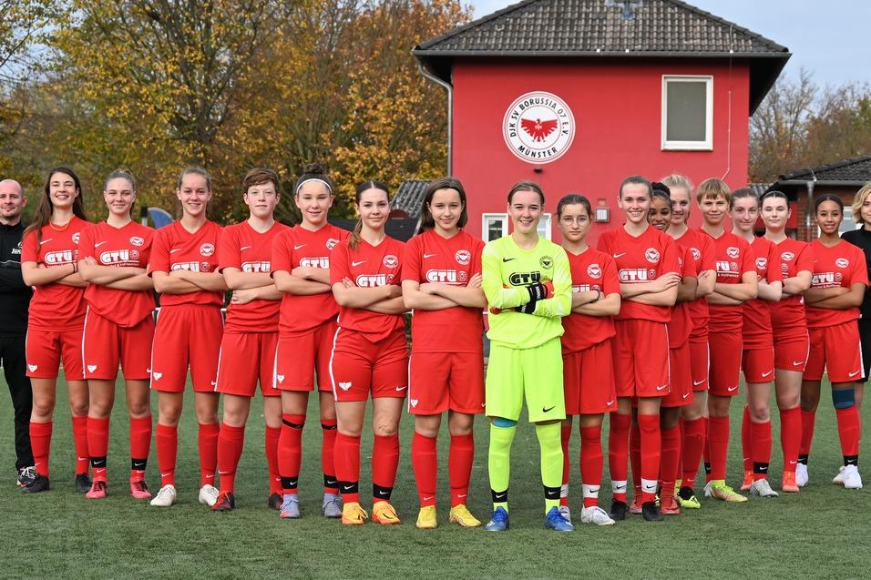 Mädchenfußballmannschaft in roten Trikots steht auf einem Sportplatz, hinter ihnen ein Vereinsgebäude mit Logo.