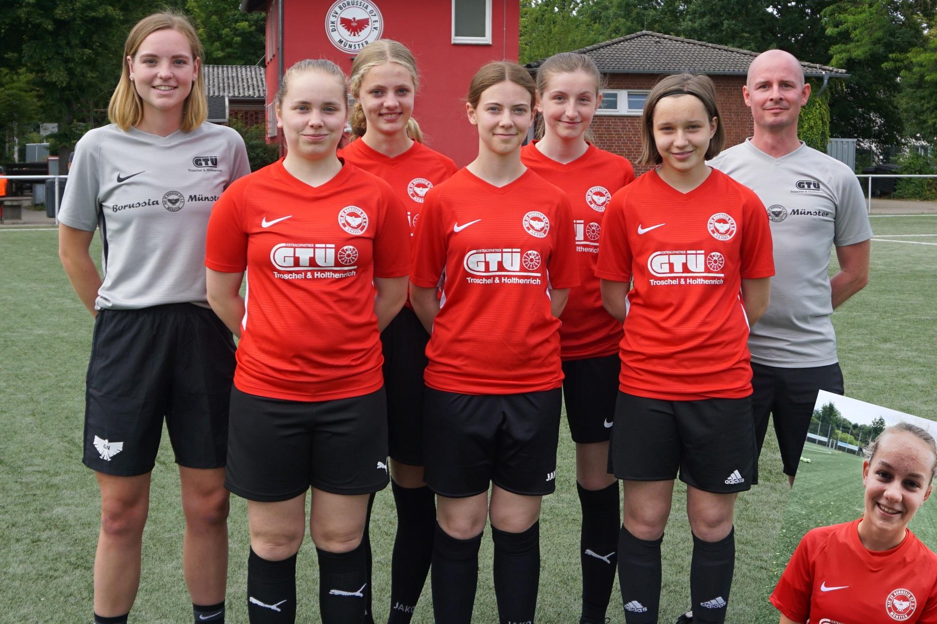 Sechsergruppe von Frauen in roten Fußballtrikots, stehend auf einem Sportplatz, mit Trainer im Hintergrund.
