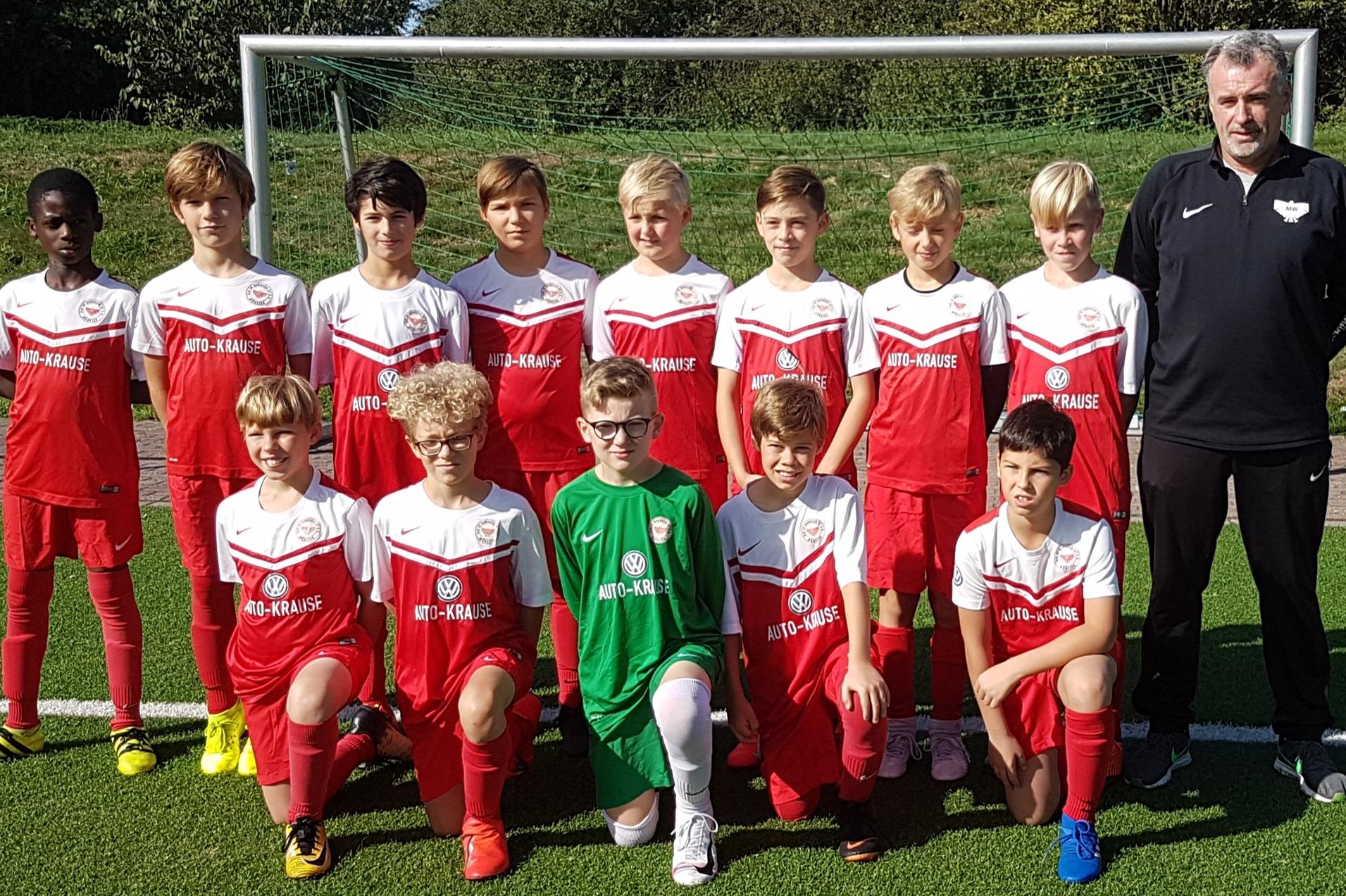Teamfoto einer Jugendfußballmannschaft in roten Trikots mit Trainer, auf einem Kunstrasenfeld hinter einem Tor.