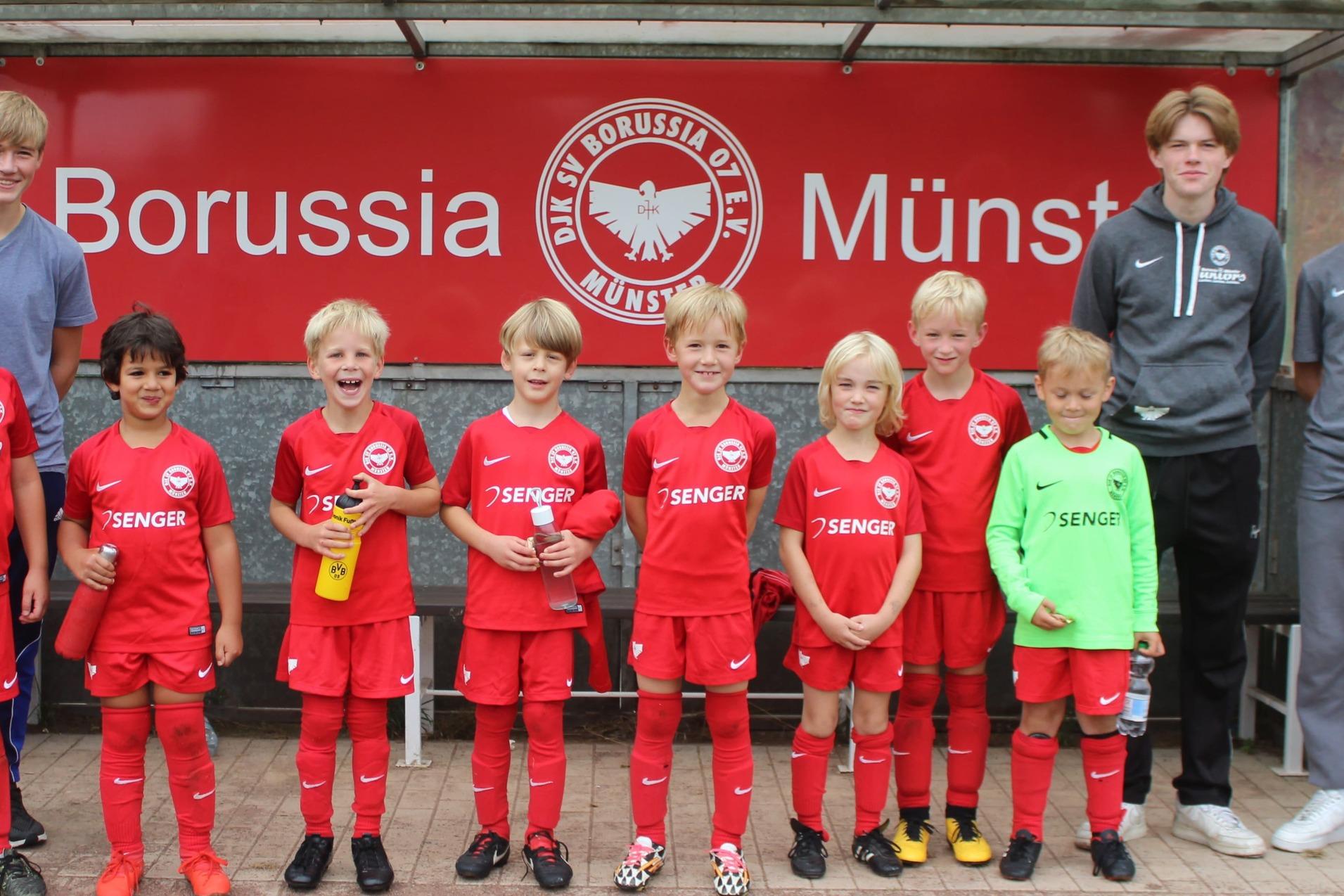 Kinderfußballmannschaft in roten Trikots steht lächelnd vor einem Vereinsbanner von Borussia Münster.