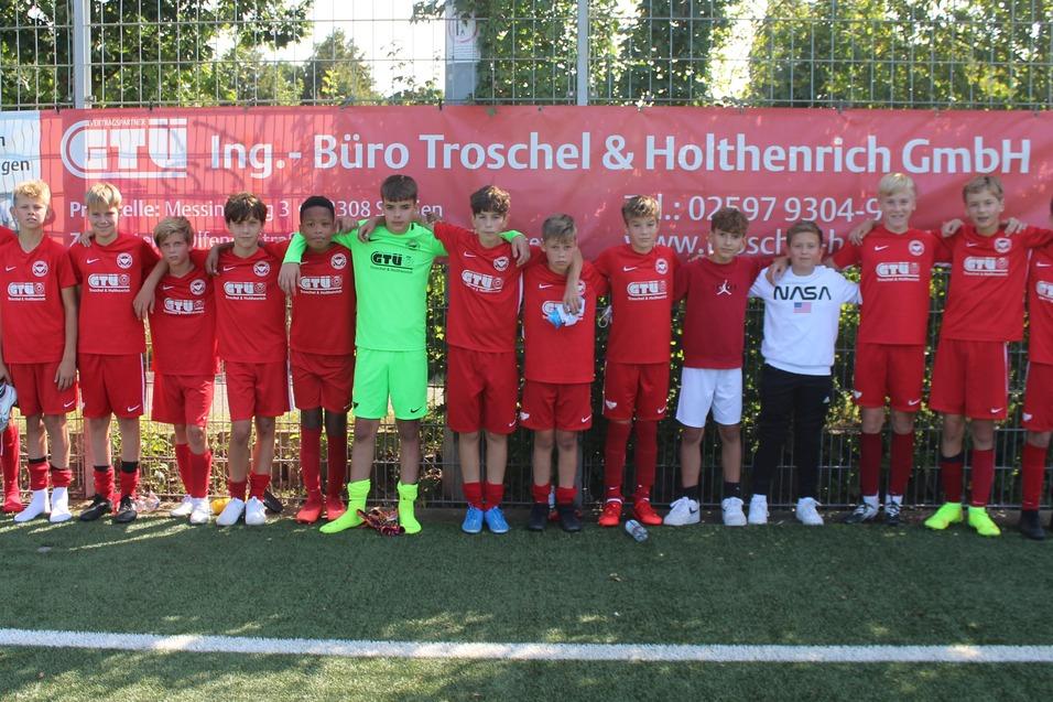Gruppe von Fußballspielern in roten Trikots posiert auf einem Sportplatz mit Werbebanner im Hintergrund.