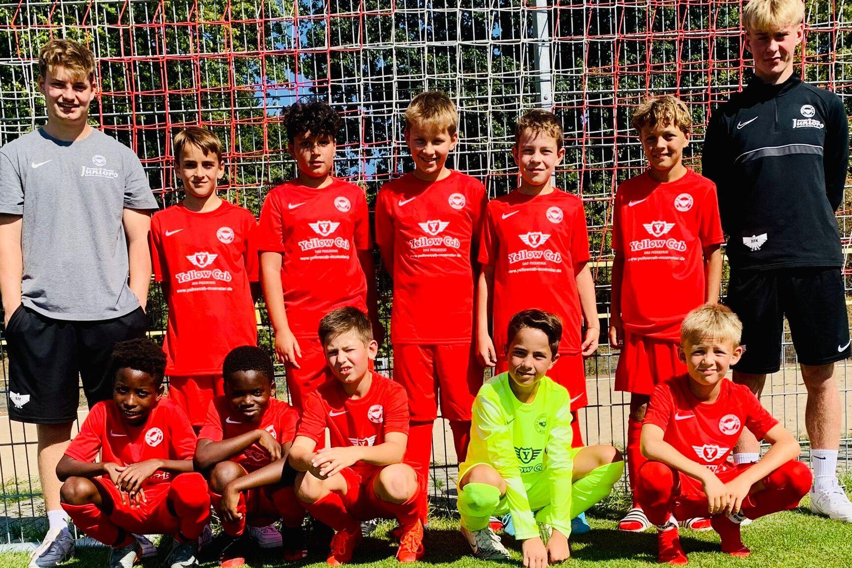 Gruppe von zehn jungen Fußballspielern in roten Trikots, posierend vor einem Fußballtor auf einem grünen Platz.