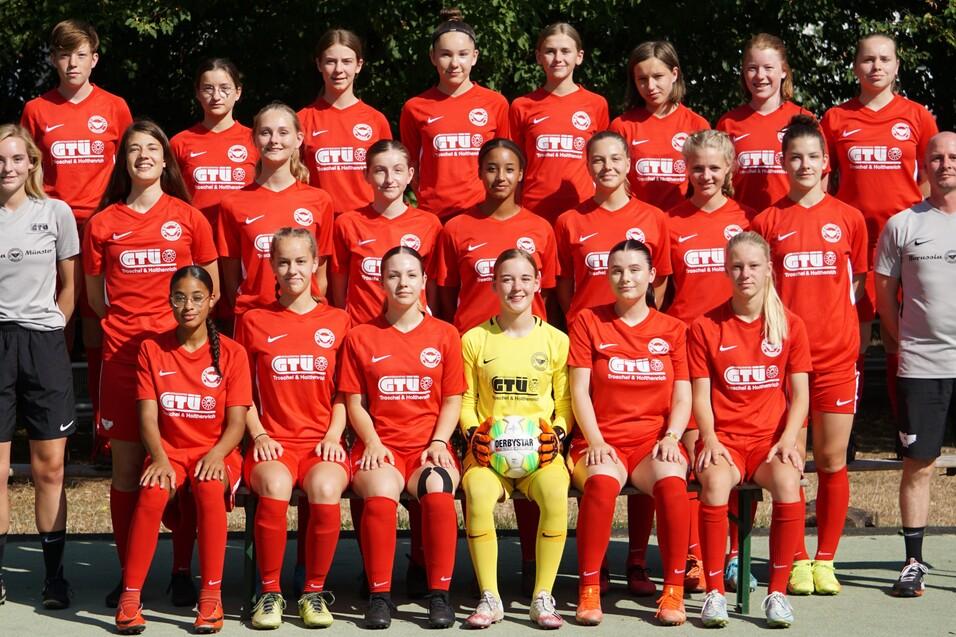 Gruppenfoto einer Fußballmannschaft in roten Trikots, inklusive Trainer, auf einem Sportplatz im Freien.