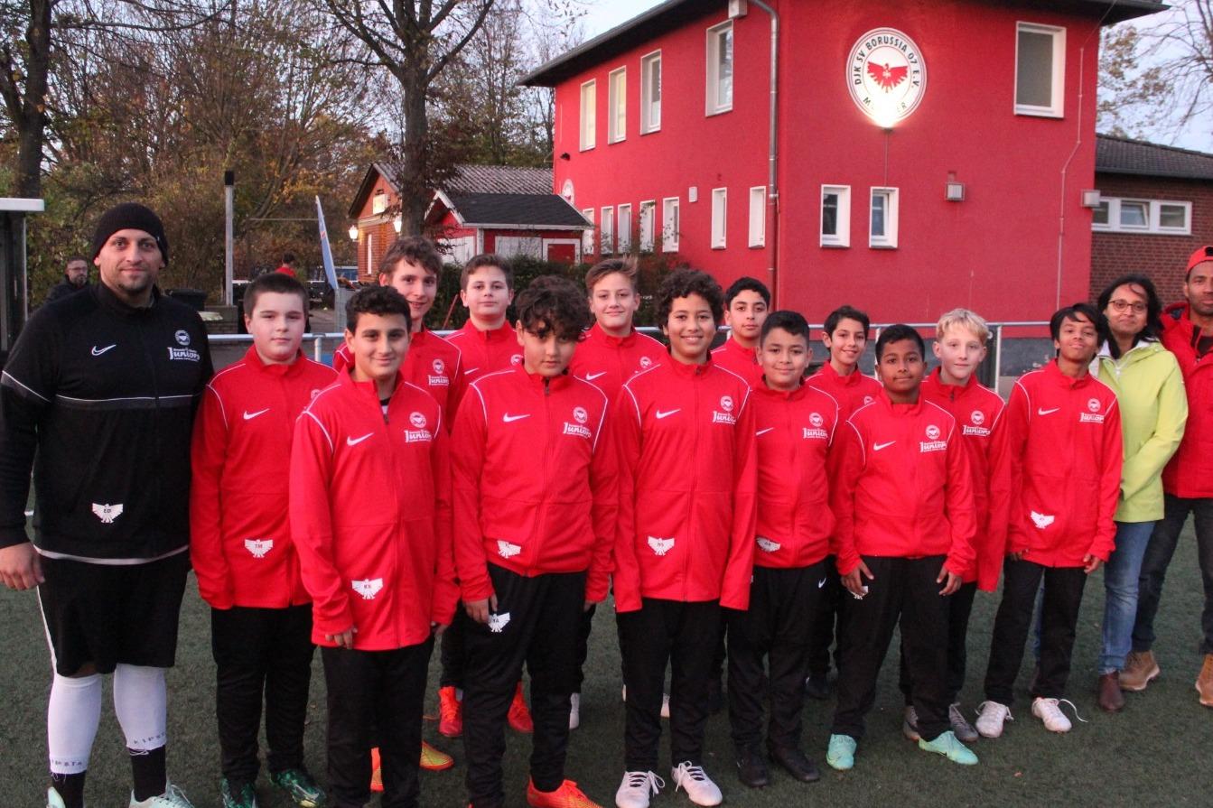 Gruppe von Fußballspielern in roten Trainingsanzügen steht vor einem Sportgebäude auf einem Kunstrasenplatz.