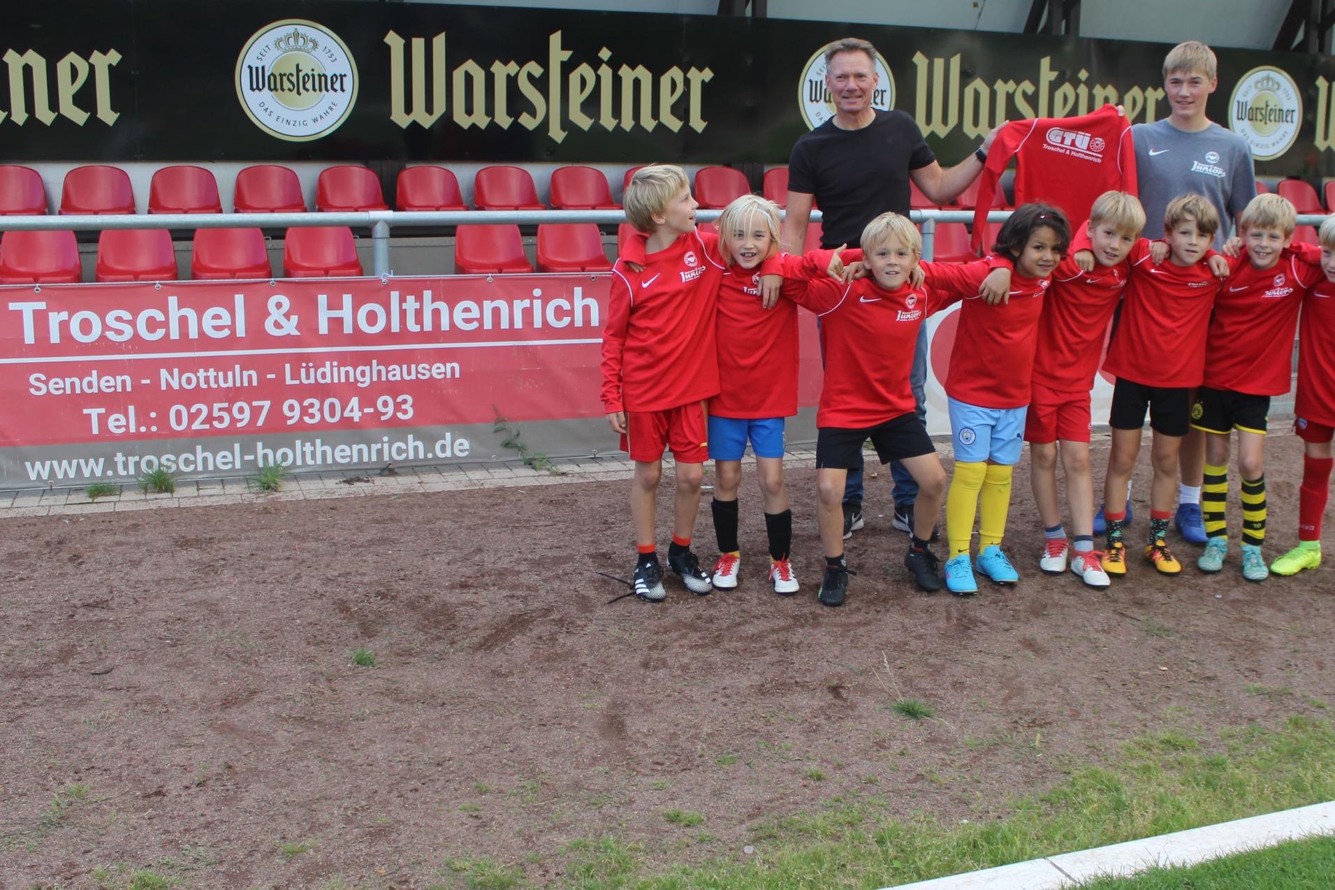 Gruppe von zehn Jungen in roten Trikots posiert auf einem Fußballplatz, Hintergrund zeigt Zuschauertribüne und Banner.