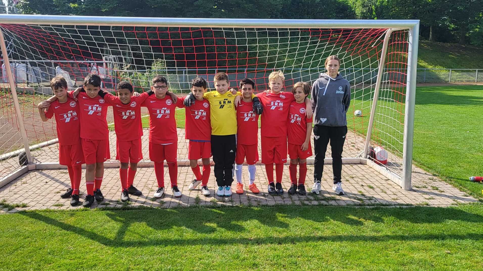 Gruppe von zehn Kindern in roten Fußballtrikots, die vor einem Tor auf dem Spielfeld stehen, begleitet von einem Trainer.