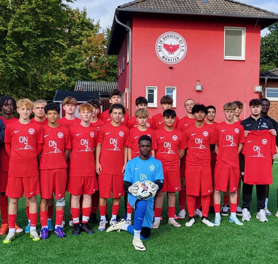 Gruppenfoto einer Jugendfußballmannschaft in roten Trikots vor einem roten Vereinsgebäude in Münster.