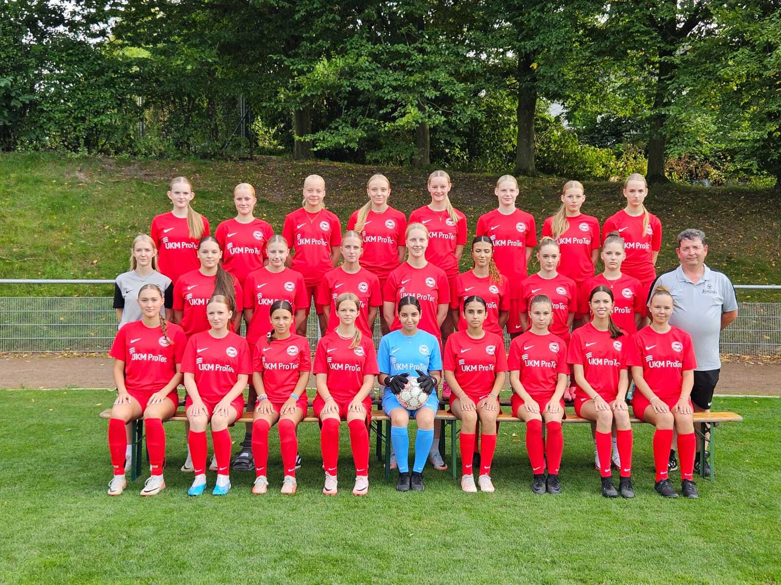 Mannschaftsfoto einer Fußballmannschaft mit 20 Spielerinnen in roten Trikots und einem Trainer auf einer Wiese.