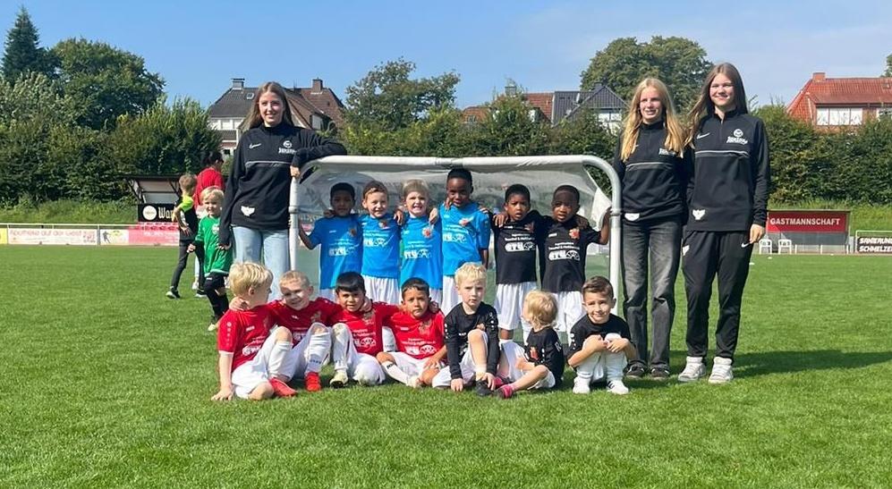 Gruppenfoto von Kindern in Fußballtrikots und zwei Trainerinnen vor einem Tor auf einem grünen Spielfeld.