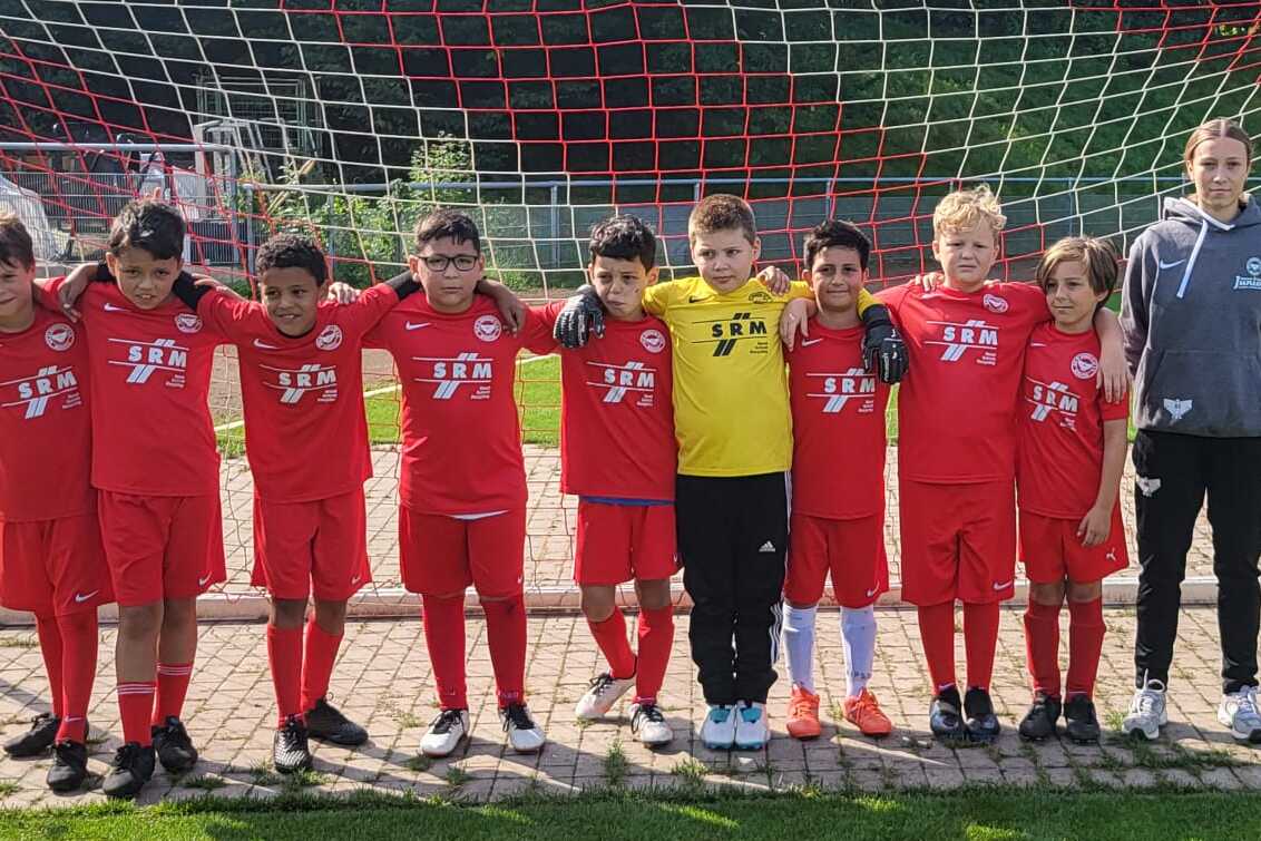 Gruppe von zehn Kindern in roten Fußballtrikots, die vor einem Tor auf dem Spielfeld stehen, begleitet von einem Trainer.