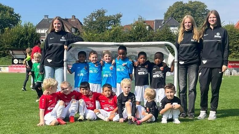 Gruppenfoto von Kindern in Fußballtrikots und zwei Trainerinnen vor einem Tor auf einem grünen Spielfeld.
