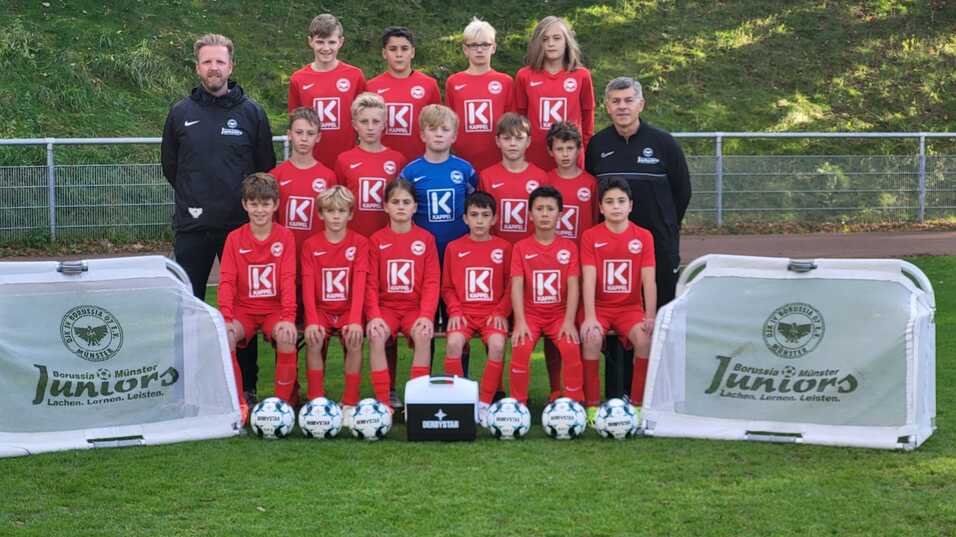 Mannschaftsbild einer Jugendfußballmannschaft in roten Trikots mit Trainern, Bällen und Werbebannern auf dem Rasen.