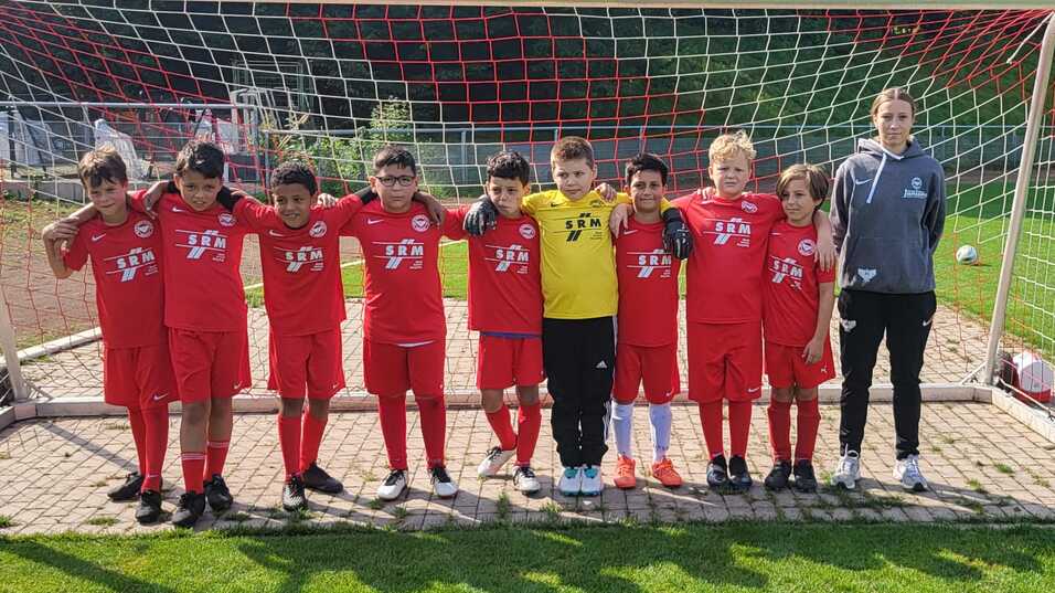 Gruppe von zehn Kindern in roten Fußballtrikots, die vor einem Tor auf dem Spielfeld stehen, begleitet von einem Trainer.