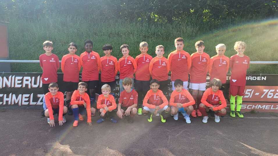 Gruppe von Jugendlichen in roten Trikots, posierend auf einem Fußballplatz vor einem Werbebanner.