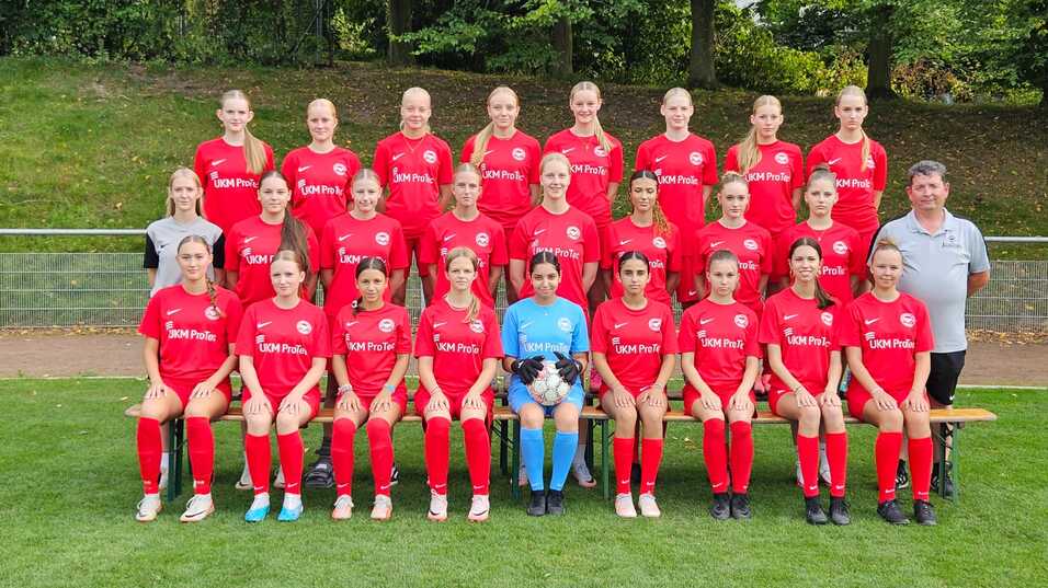 Mannschaftsfoto einer Fußballmannschaft mit 20 Spielerinnen in roten Trikots und einem Trainer auf einer Wiese.