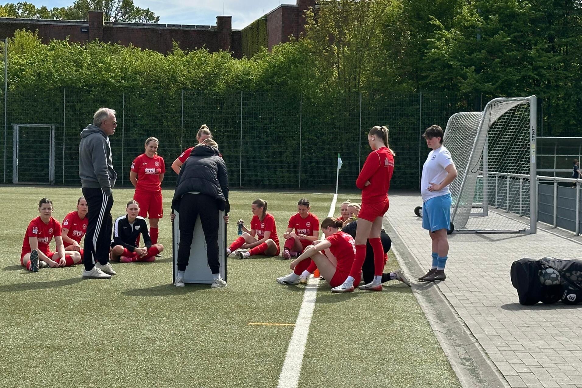 Trainer spricht mit einer Gruppe Fußballspielerinnen in roten Trikots auf einem Kunstrasenplatz.