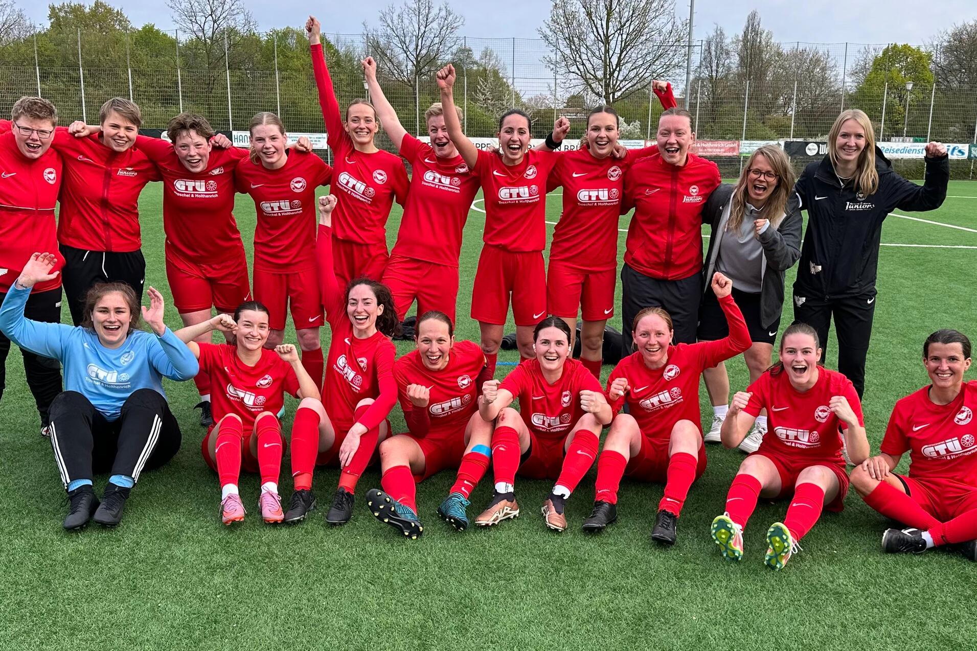 Gruppenselfie von 18 Frauen in roten Fußballtrikots, die auf einem Platz jubeln und die Arme in die Luft strecken.