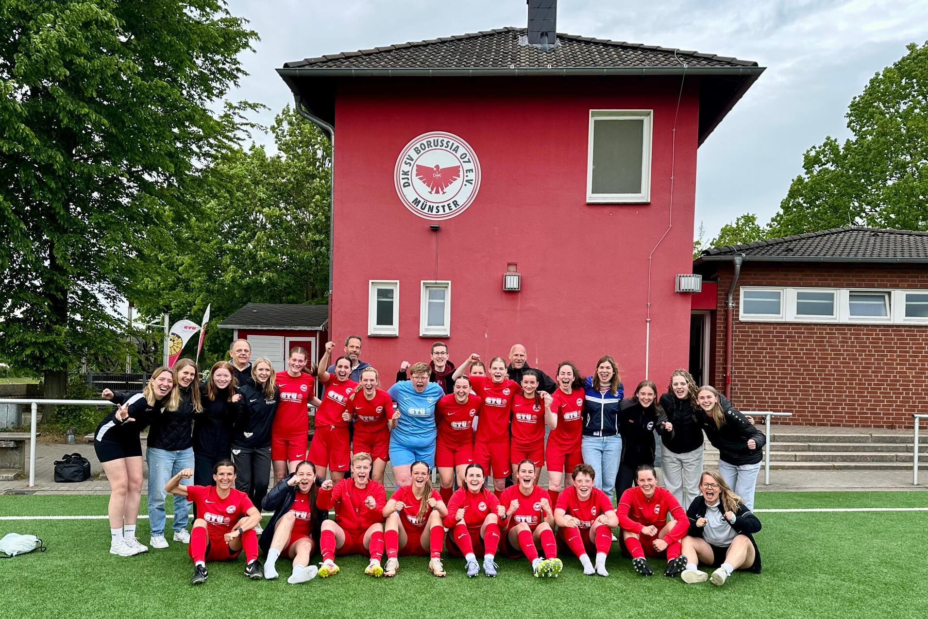 Gruppenfoto einer Fußballmannschaft in roten Trikots vor einem roten Vereinsgebäude, umgeben von Bäumen.