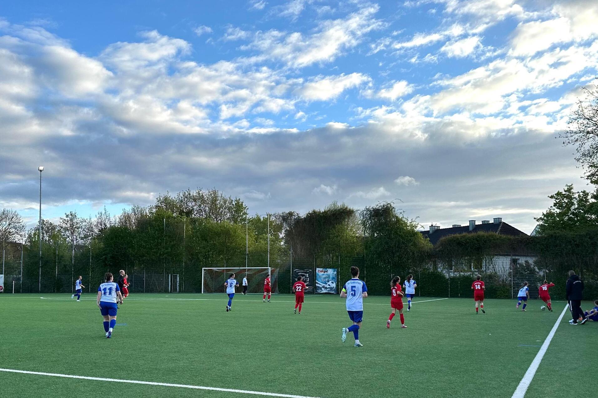 Fußballspiel auf einem Kunstrasenplatz, Spieler in roten und blauen Trikots, Bäume und Wolken im Hintergrund.