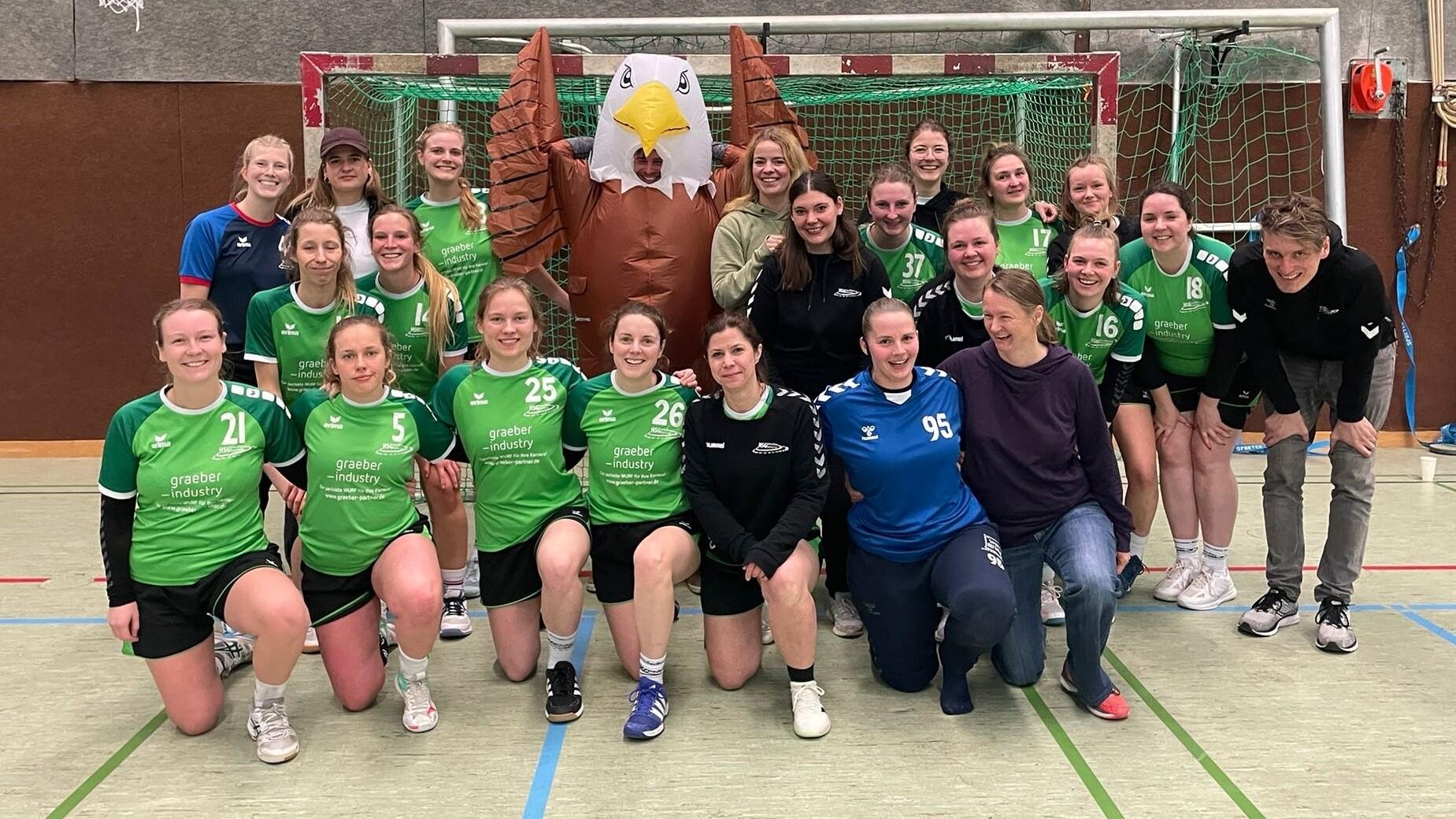 Gruppe von Handballspielerinnen in grünen Trikots posiert vor einem Tor mit einem großen Adler-Banner.
