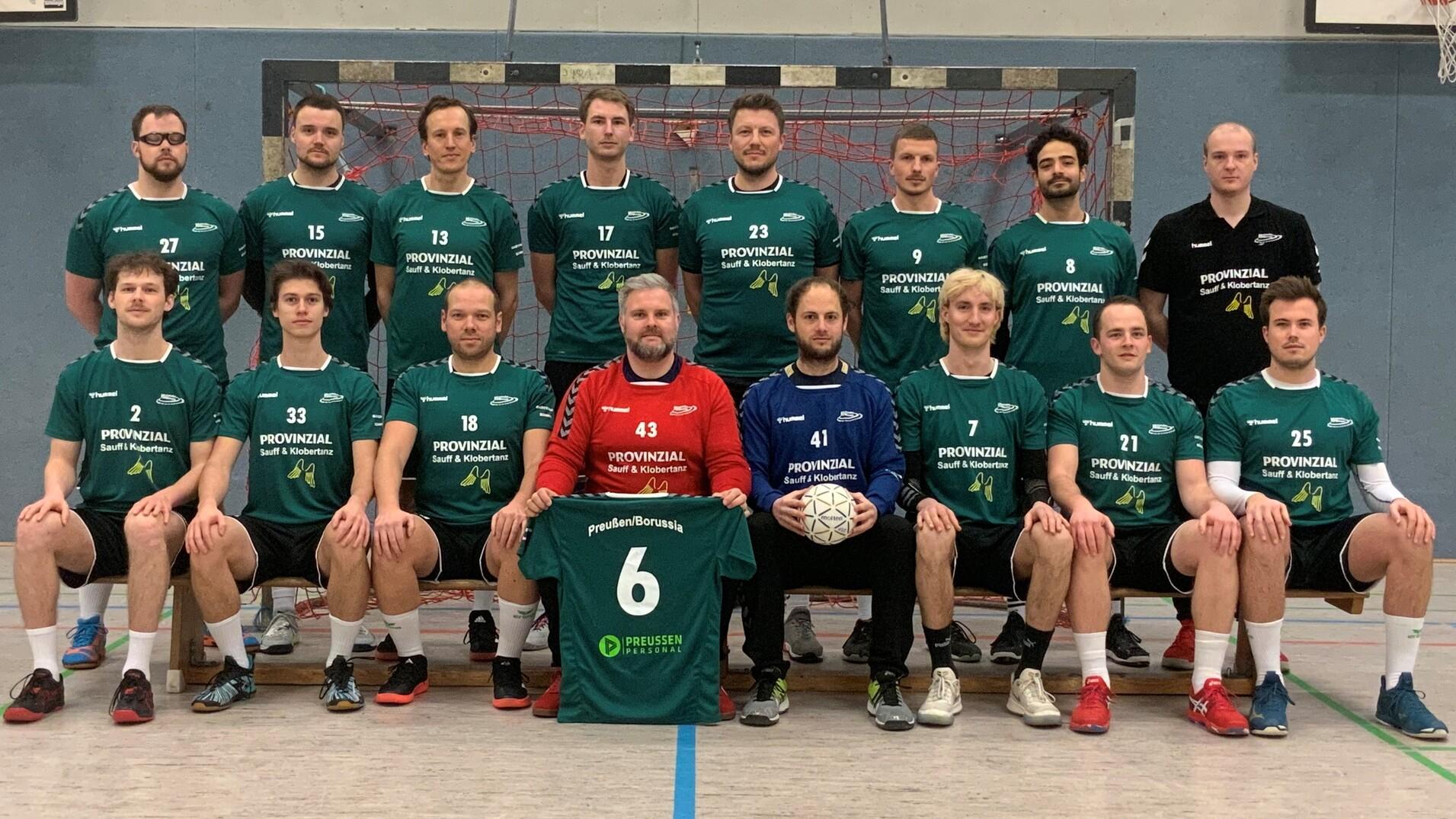 Mannschaftsfoto einer Handballmannschaft in grünen Trikots, aufgestellt vor einem Handballtor in einer Sporthalle.