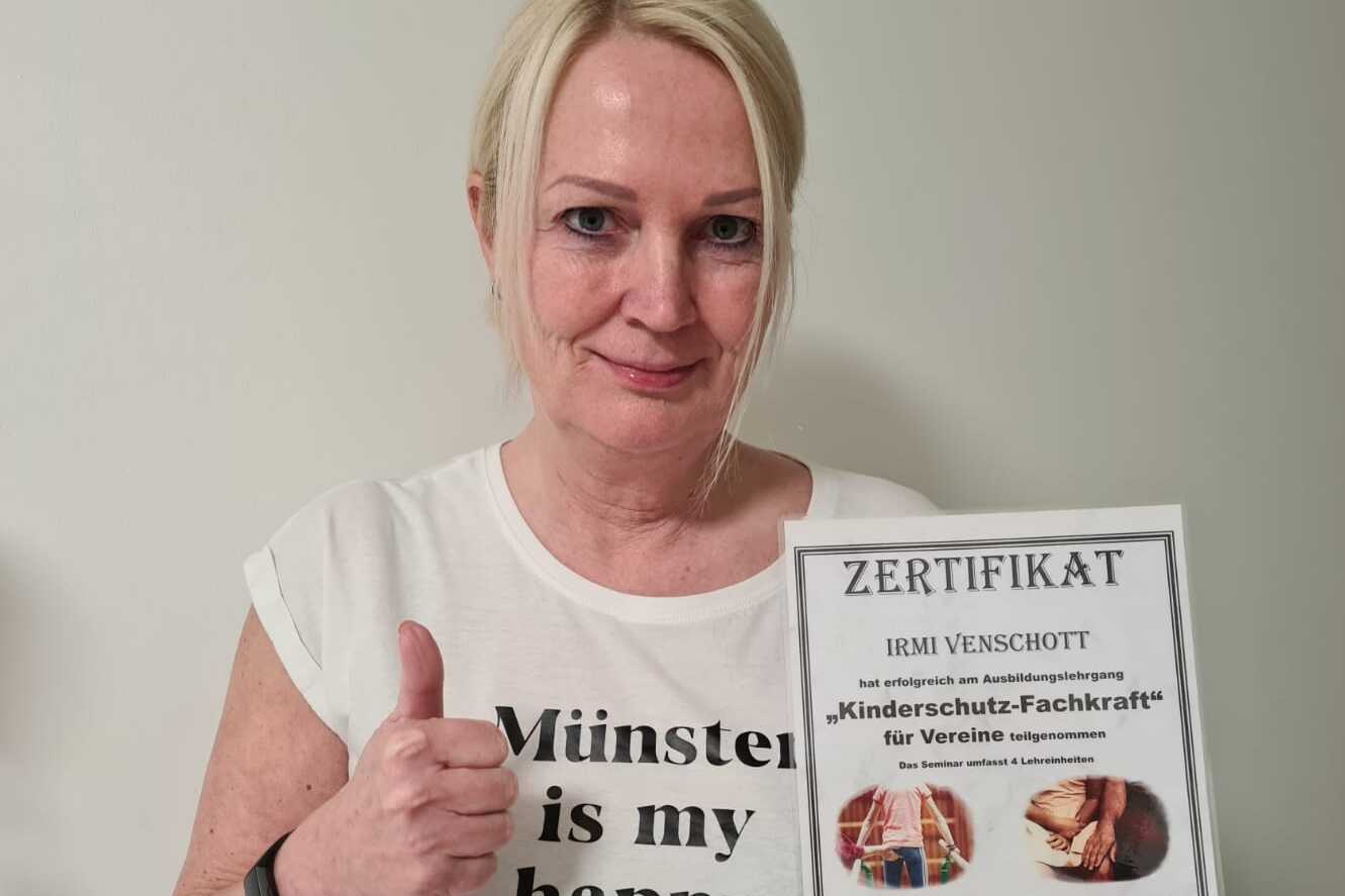 Frau mit Daumen hoch präsentiert ein Zertifikat über "Kinderschutz-Fachkraft" vor einer weißen Wand.