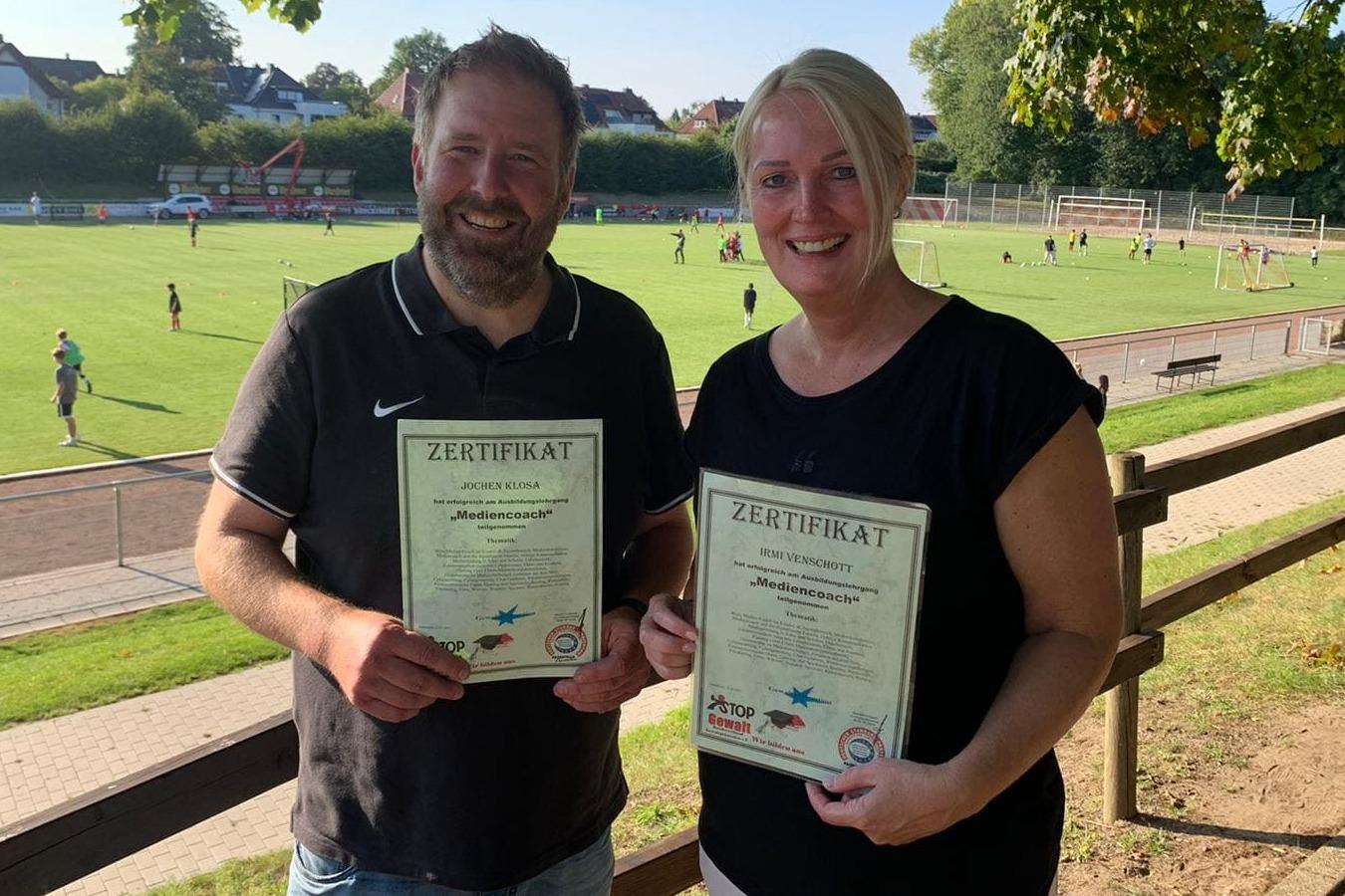 Zwei Personen halten stolz ihre Zertifikate vor einem Sportplatz mit Fußballspielern im Hintergrund.