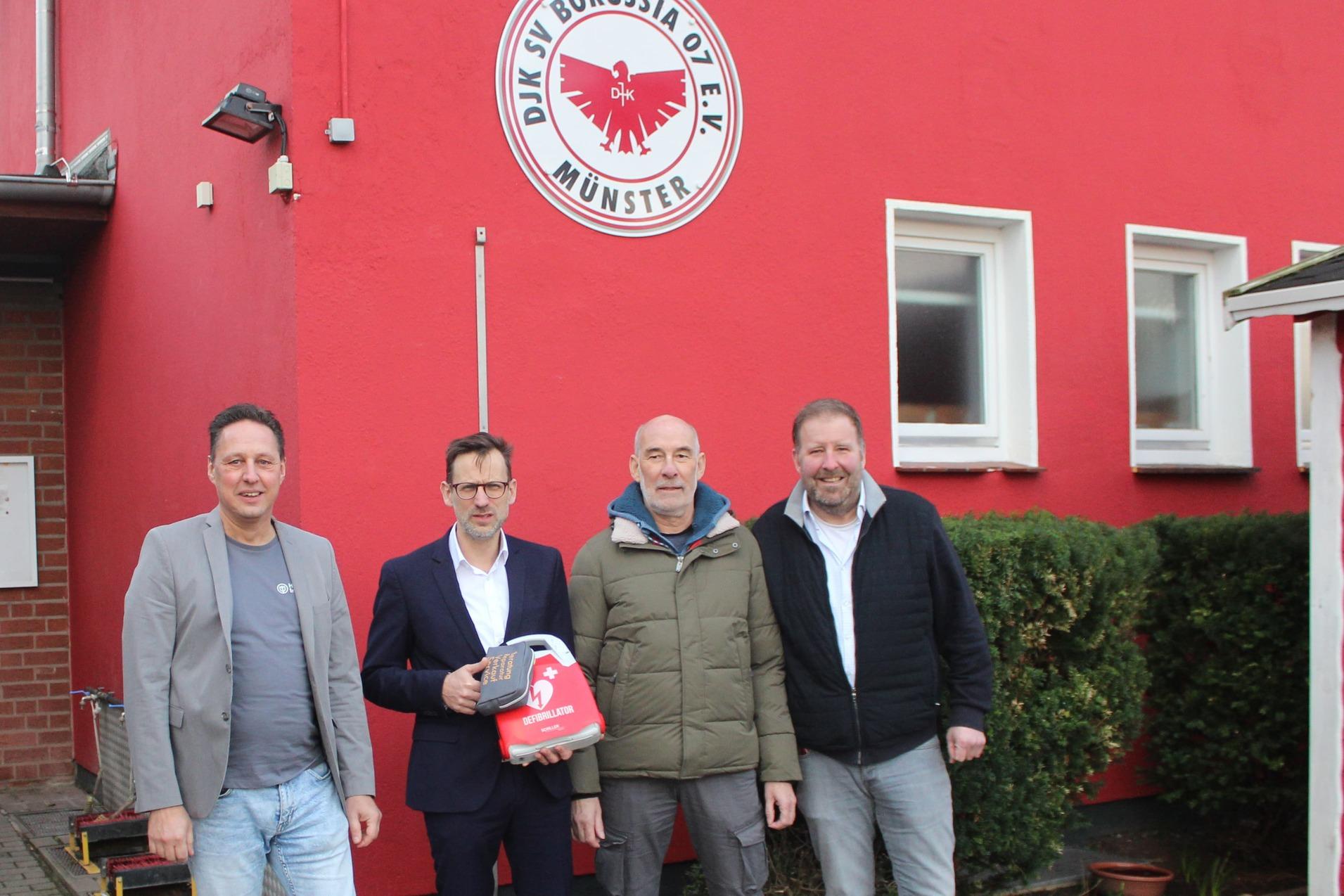 Vier Männer stehen vor einem roten Gebäude mit dem Logo von Borussia Münster, während sie Fahnen in den Händen halten.