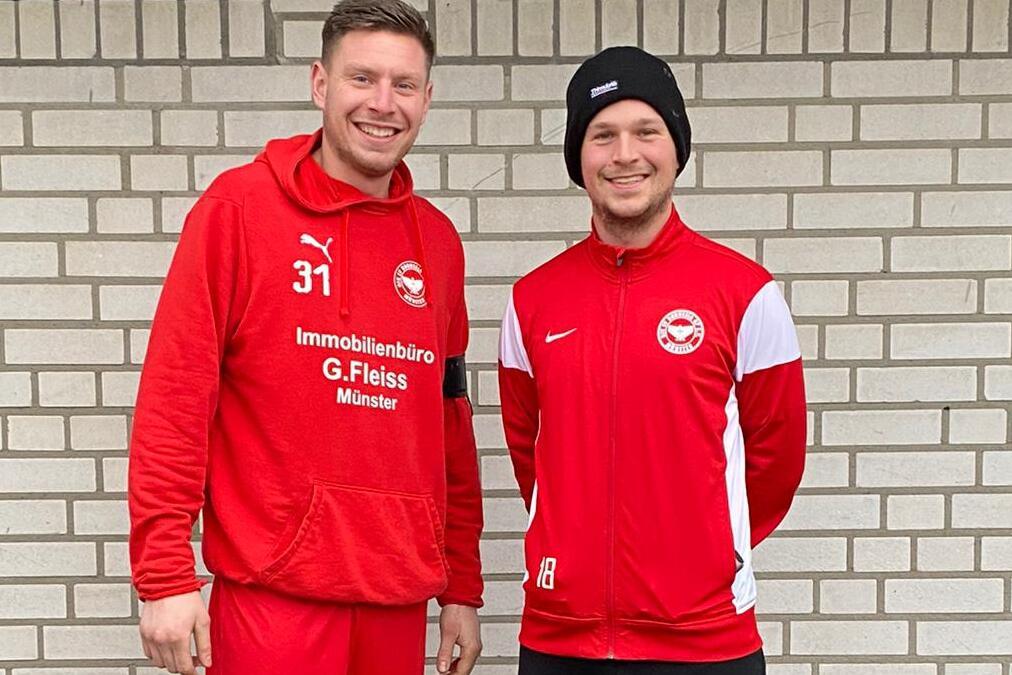 Zwei Fußballspieler in roter Sportkleidung stehen vor einer gemusterten Backsteinwand.