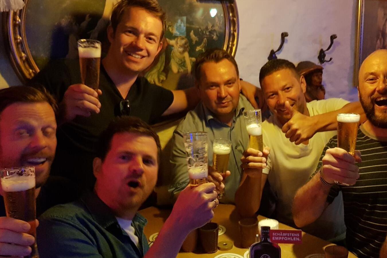 Gruppierung von sieben Männern, die an einem Tisch in einer Bar Biergläser heben und lächeln, umgeben von Wänden und Dekor.