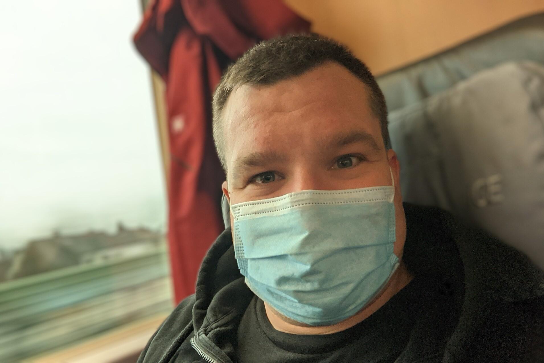 Mann mit Mundschutz sitzt im Zug und lächelt, Hintergrund zeigt verschwommene Landschaft.