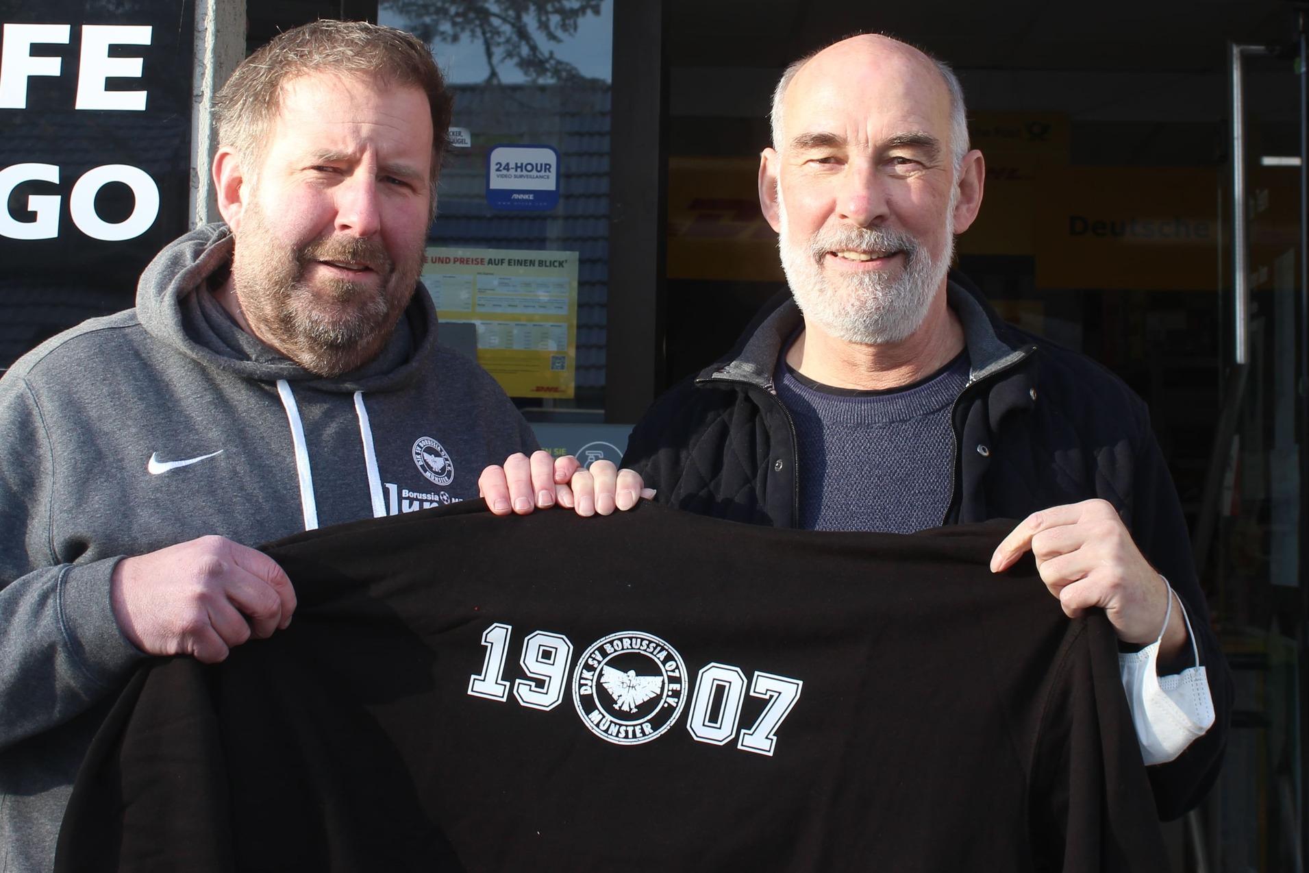 Zwei Männer stehen vor einem Café und halten ein schwarzes T-Shirt mit der Aufschrift „19007“. Im Hintergrund ist das Café-Schild sichtbar.