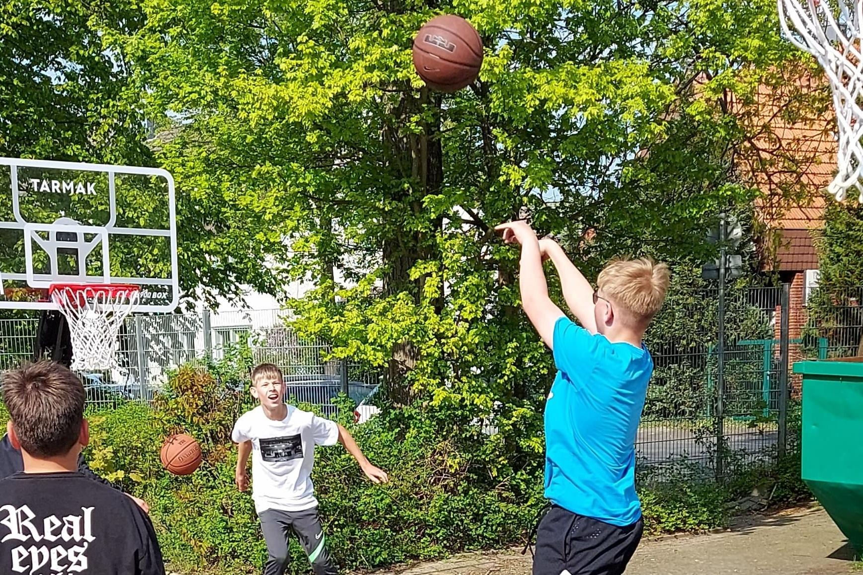 Junger Spieler wirft Basketball zum Korb, während andere Spieler im Hintergrund warten, auf einem Freiplatz.
