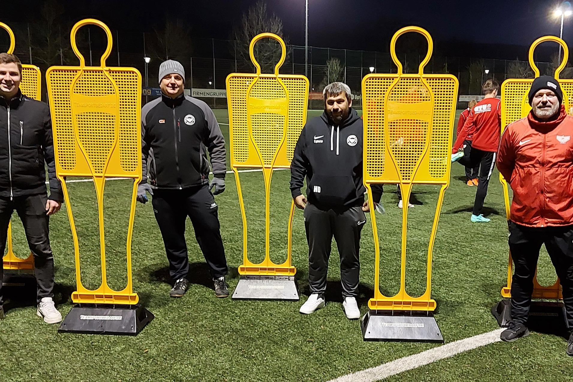 Training auf dem Fußballplatz mit vier orangefarbenen Trainingshilfen und drei Trainern in Sportkleidung.